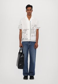 Camisa blanca de manga corta con dibujos en línea negra de una casa y árboles, combinada con unos jeans azules y una bolsa tote negra. Diseño sencillo, corte relajado.