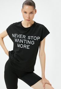 Sort T-shirt med korte ærmer, der har fed hvid tekst "NEVER STOP WANTING MORE." Sammen med sorte cykelshorts skaber det et sammenhængende atletisk look.