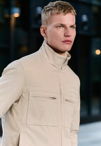Beige zip-up jack met staande kraag, voorzien van twee voorste met rits afgesloten zakken en een gladde, lichte stof. Duidelijke stikseldetails zichtbaar.