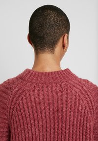 Pull en maille de couleur rose poussiéreux, doux, avec une texture côtelée. Présente un col arrondi et une coupe décontractée, montrant la vue arrière.