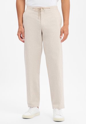 Uomo che indossa pantaloni di lino beige con coulisse in vita e scarpe da ginnastica bianche, in piedi contro uno sfondo bianco semplice.