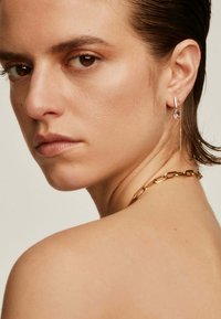 Collar de cadena de oro con grandes eslabones; los pendientes colgantes presentan una piedra preciosa rosa y piedras de acento, todo sobre un fondo neutral.