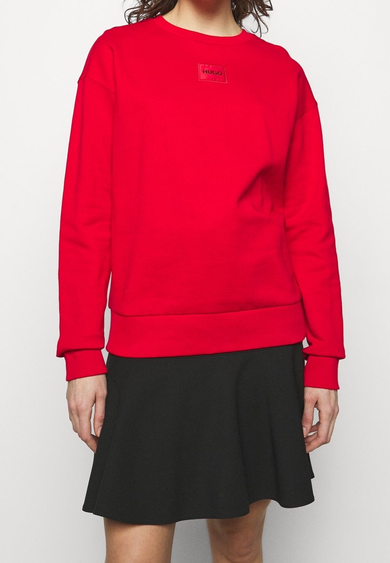 Femme portant un sweat-shirt à manches longues rouge vif avec un petit logo "HUGO" sur la poitrine et une jupe évasée noire longueur genou.