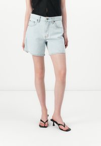 Pantalones cortos de denim azul claro con cintura alta, detalles de bolsillos y dobladillo deshilachado. Combinados con sandalias negras de tacón y una blusa negra.