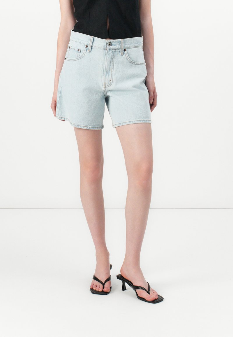 Pantalones cortos de denim azul claro con cintura alta, detalles de bolsillos y dobladillo deshilachado. Combinados con sandalias negras de tacón y una blusa negra.