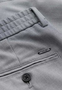 Gros plan d'un pantalon en tissu gris texturé montrant une boucle de ceinture, une poche cousue avec un bouton, et une petite plaque métallique indiquant « ombre dept. since 1985. »
