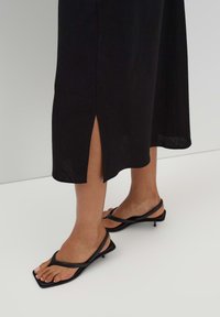 Schwarzes Midikleid mit einem seitlichen Schlitz, aus leichtem Stoff gefertigt. Kombiniert mit schwarzen Sandalen mit Riemchen, die eine quadratische Zehenpartie und einen Absatz haben.