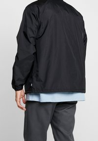 Giacca nera leggera con una texture liscia, polsini elasticizzati e orlo dritto, indossata sopra una camicia azzurro chiaro e pantaloni grigi.