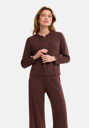 Kobieta ubrana w brązową bluzę z kapturem na zamek i sznurki oraz dopasowane szerokie spodnie, stojąca na jasnym, jednolitym tle.