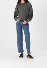 Felpa grigia con stampa paisley nera a rilievo sulle maniche, abbinata a jeans dritti blu e sneakers nere con strisce bianche.