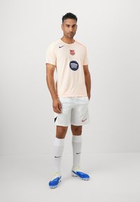 Man standing wearing light peach FC Barcelona soccer jersey, white shorts, white socks with black Nike logos, and blue and white cleats against white background.