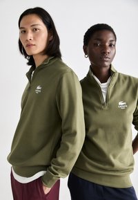Duomenų dvi asmenys dėvi žalius užtraukiamus megztinius su baltais Lacoste logotipais. Megztiniai turi minkštą tekstūrą ir aukštą apykaklę.