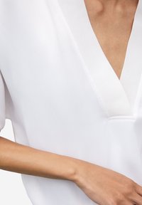 Blouse blanche avec un profond décolleté en V orné d'une bordure texturée. Manches courtes et amples. Tissu lisse avec un léger éclat et design minimaliste.