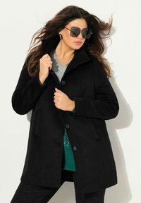 Cappotto nero in misto lana con collo alto, chiusura a bottoni e due tasche laterali, indossato sopra un top grigio e un maglione verde. Occhiali scuri completano il look.