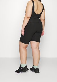 Kurvig person klädd i svart ärmlös topp och högmidjade svarta cykelshorts, kombinerat med svarta sneakers och ljusgröna strumpor, stående mot vit vägg.