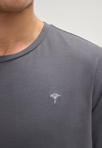 Grå kortærmet T-shirt lavet af blødt stof, med et lille hvidt broderet logo på venstre brystområde. Rund halsudskæring, minimalistisk design.