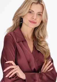 Cămașă din satin burgundy, cu guler și manșete cu nasturi, având o textură fină. Părul lung și cerceii lungi completează aspectul.