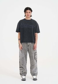 Czarny, oversize t-shirt z denimu z nadrukiem "Ed Hardy 1971" w kolorze szarym. Szare spodnie cargo z białą haftowaną aplikacją i wieloma kieszeniami. Czarno-białe buty.