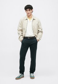 Beige bomber dzseki egy fehér pulóver felett, sötétzöld nadrág és zöld sportcipő fehér fűzősávval. Simább textúrák és egyszerű dizájn.