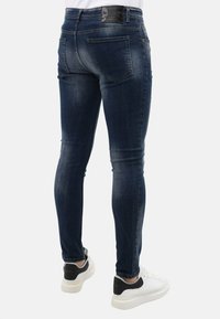 Strakke jeans in donkerblauwe denim, met een klassieke vijf-pocketontwerp en een zwart logo-embleem op de tailleband. Gecombineerd met witte sneakers.