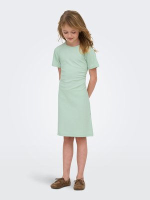 Ung pige med lysebrunt hår iført en lysegrøn kjole med korte ærmer, der går til knæene, og brune sko, kigger ned med hænderne på ryggen.