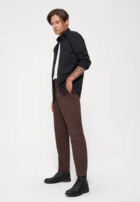 Camisa negra de botones, camiseta blanca debajo, pantalones marrones y botas negras con cordones. Tela suave con un ajuste ceñido.