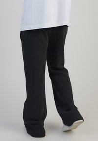 Melni sporta bikses ar brīvu piegriezumu, taisnām kājām un ribotām piedurknēm, kopā ar baltu T-kreklu un baltiem sporta apaviem uz vienkrāsaina fona.