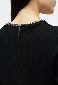 Γυναίκα που φοράει μαύρο ριμπ πουλόβερ με ασημένια αλυσίδα κατά μήκος του πίσω μέρους του λαιμού και μια μικρή ετικέτα σύρτη φερμουάρ.