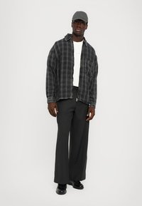 Camicia a quadri nera e grigia, t-shirt a girocollo bianca, pantaloni neri a vita alta, cappellino nero e scarpe. Il modello è in piedi contro uno sfondo neutro.