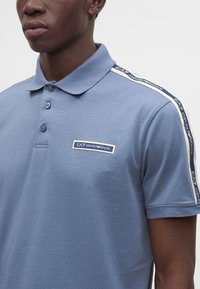Άνδρας που φοράει μπλε πόλο EA7 Emporio Armani με κουμπωτό γιακά και κορδέλα με λογότυπο κατά μήκος της ραφής στον ώμο.