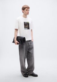 Λευκό μπλουζάκι με μαύρο γραφικό τύπωμα, συνδυασμένο με άνετο γκρι τζιν. Κρατάει μαύρο τσαντάκι χειρός. Μαύρα παπούτσια. Απλός σχεδιασμός.