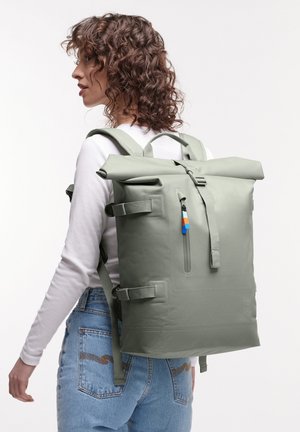 Frau mit lockigem Haar, die ein weißes Langarmshirt und blaue Jeans trägt, trägt einen großen grauen Roll-Top-Rucksack und schaut nach links.