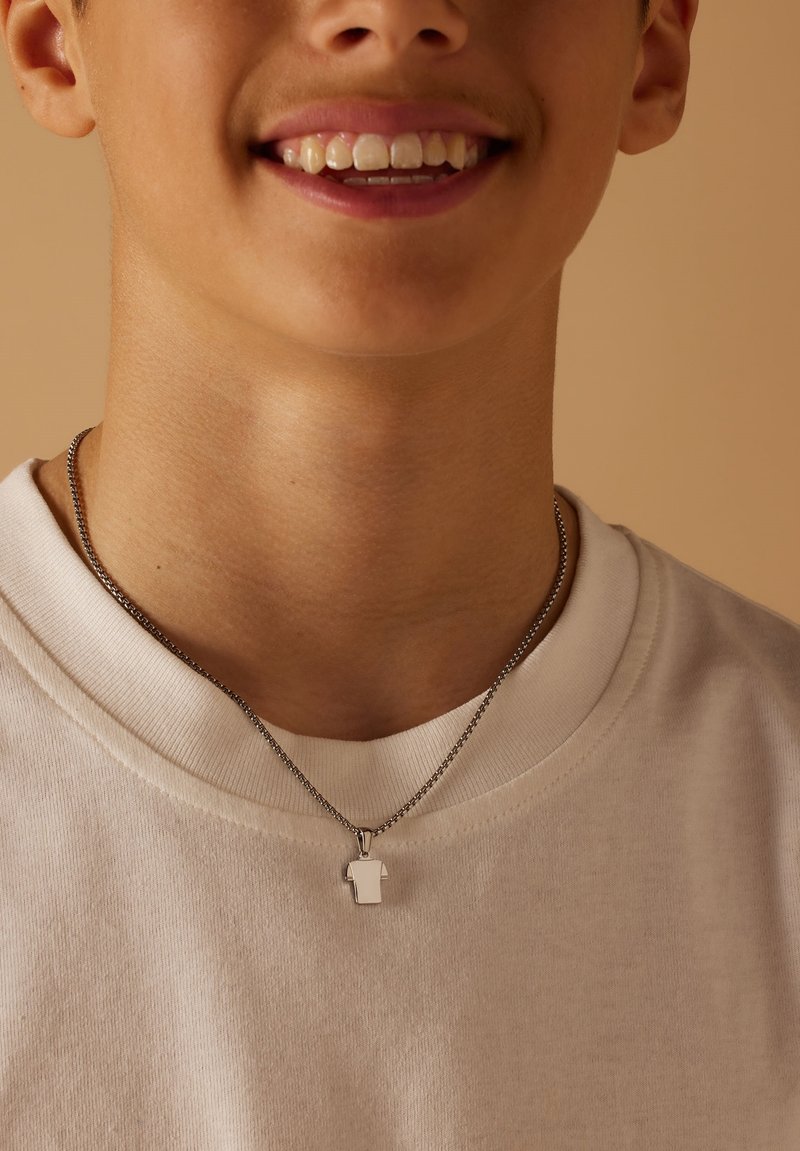 Zilverkleurige ketting met een klein, abstract hangertje in de vorm van een shirt, gedragen door een persoon in een lichte T-shirt.
