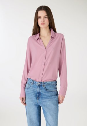 Młoda kobieta ubrana w różową, zapinaną na guziki bluzkę z długim rękawem, wpuszczoną w wysokowalne, jasnoniebieskie dżinsy, stoi na jednolitym białym tle.