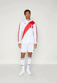 Bela dolga majica za nogomet s rdečim diagonalnim trakom, rdečimi akcenti in tremi rdečimi črtami na rokavih. Model nosi bele kratke hlače in nogometne copate.