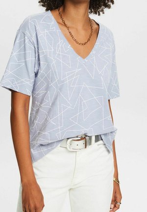 Kobieta w jasnoniebieskiej koszulce z dekoltem w kształcie litery V ozdobionej białymi geometrycznymi liniami, włożonej w białe spodnie z białym paskiem oraz z złotym łańcuszkiem na szyi.