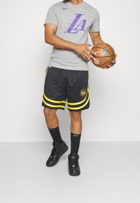 Chemise grise à manches courtes avec logo "LA" violet, short de basket noir avec des bandes jaunes, chaussettes noires et baskets noires, tenant un ballon de basket.