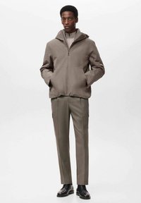 Chaqueta marrón con cremallera y capucha, combinada con pantalones beige de pierna recta. Tejido suave, diseño minimalista, cuenta con bolsillos laterales y zapatos negros.