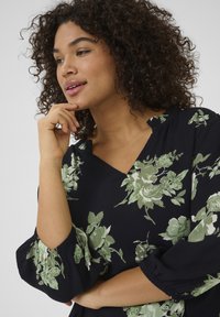 Zwarte bloemenblouse met groen botanisch patroon, V-hals en pofmouwen met elastische manchetten. Getextureerde stof en losse pasvorm.