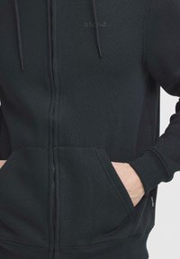 Sweat à capuche noir avec une fermeture éclair frontale, une poche kangourou et un logo discret. Le matériau semble doux avec une texture lisse et des poignets ajustés.