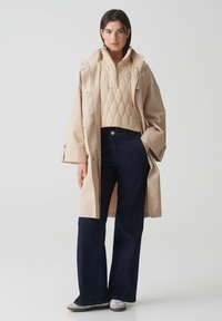 Trench coat impermeabile beige sopra un pullover trapuntato crema, abbinato a jeans a gamba larga blu scuro e scarpe slip-on grigie.