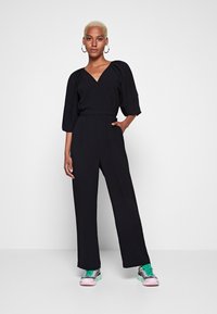 Combinaison noire avec un décolleté en V profond, des manches larges et une taille ceinturée, associée à des baskets colorées avec un mélange d'accents roses et verts.