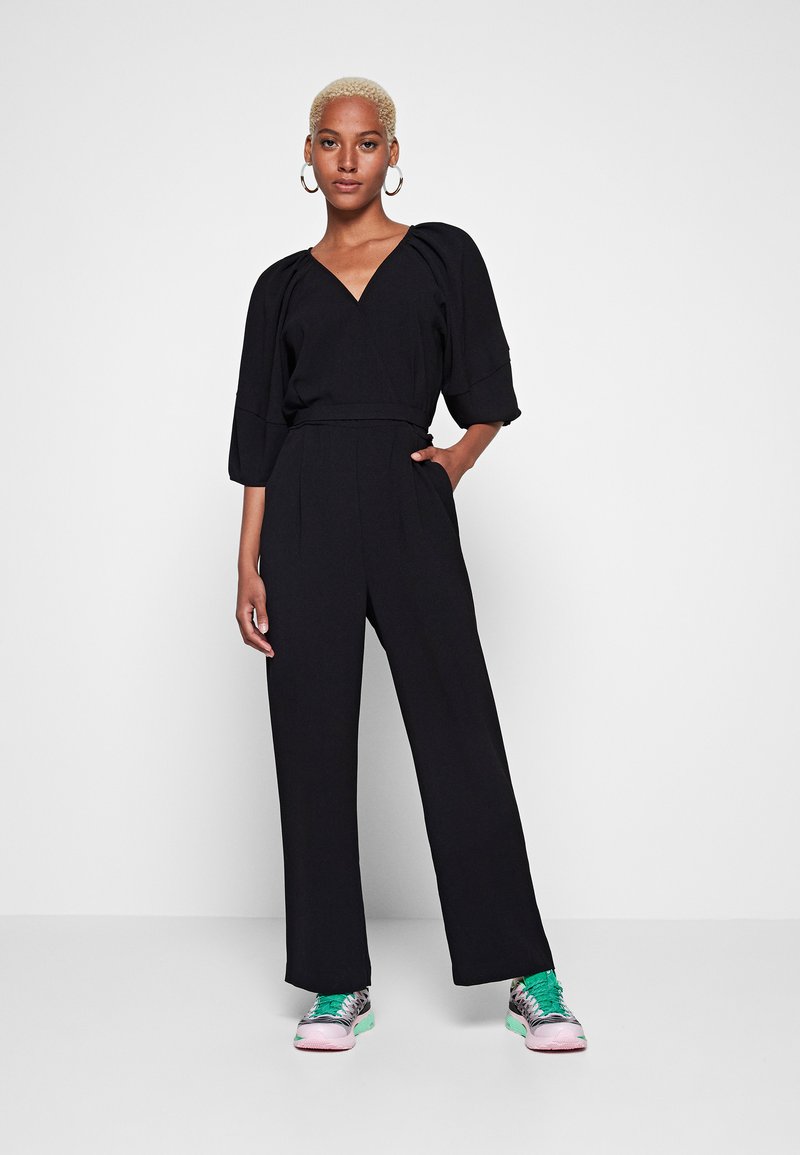Combinaison noire avec un décolleté en V profond, des manches larges et une taille ceinturée, associée à des baskets colorées avec un mélange d'accents roses et verts.