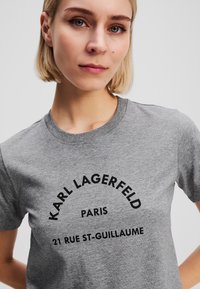 Pilkas medvilninis marškinėlis trumpomis rankovėmis ir apvalia kaklo išpjova; turi juodą tekstą: "Karl Lagerfeld Paris 21 Rue St-Guillaume."