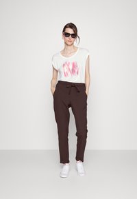 Camiseta branca com estampa abstrata em rosa, calças castanhas ajustadas e tênis brancos. Os acessórios incluem óculos de sol pretos e brincos de argola.
