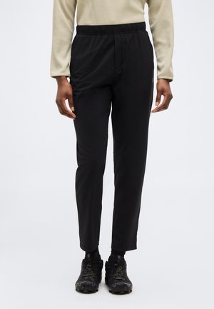 Άτομο που φοράει μαύρο παντελόνι με στενή εφαρμογή, μαύρα αθλητικά παπούτσια με κορδόνια, μπεζ μακρυμάνικη κορυφή με υφή, στέκεται με τα χέρια χαλαρά δίπλα στο σώμα.