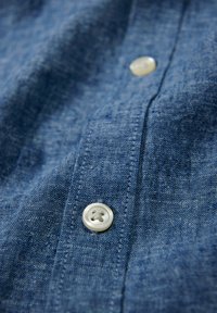 Blauwe chambray stof met een textuurafwerking, met een witte knop met vier gaten en een net gestikte lijst.