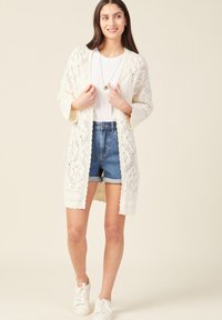 Biały kardigan szydełkowy z otwartym przodem, ze skomplikowanymi wzorami, półdługimi rękawami, zestawiony z białą bluzką i dżinsowymi szortami.
