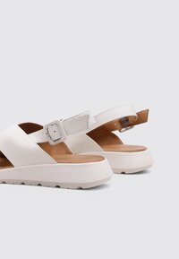 Sandalias blancas con suela plana, diseño de tiras cruzadas y hebilla ajustable. Parte superior de piel suave con un acento de talón texturizado y plantilla de madera.