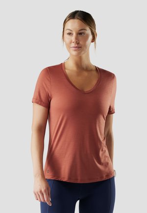 Mujer de piel clara y cabello recogido, vestida con una camiseta de manga corta color óxido con cuello en V y leggings azul oscuro, de pie frente a un fondo gris.